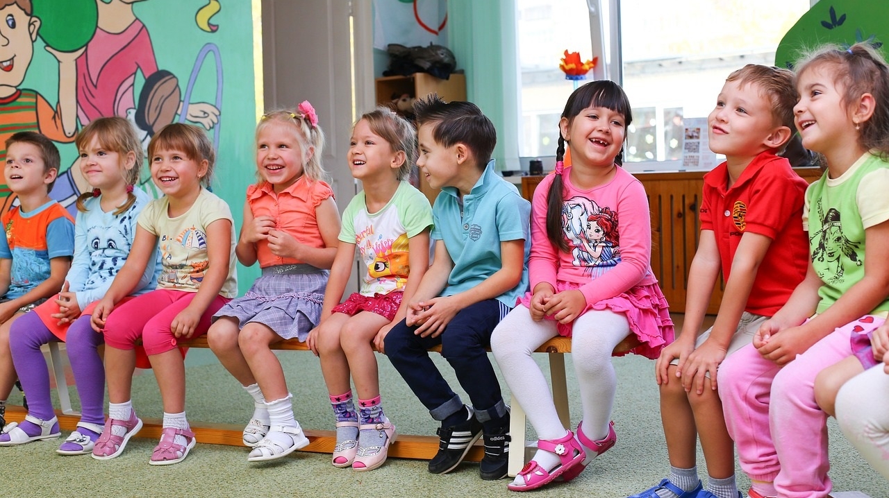 Eine Gruppe verschiedener Kinder, die im Kindergarten auf einer Bank sitzt, die Stimmung ist fröhlich, die Kinder lachen