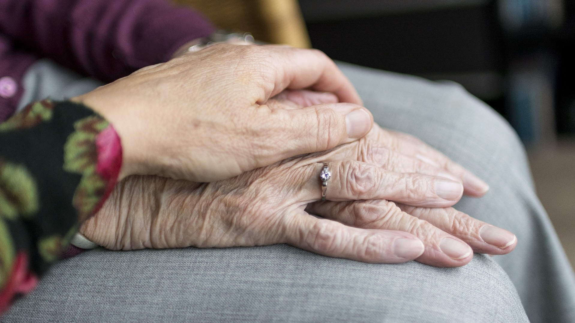eine ältere Dame hat die Hände auf ihrem Schoß übereinander gefaltet liegen, die Hand einer weiteren Frau liegt beschützend über ihren Händen