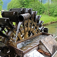 Altes Holzmühlenrad mit Wasserschöpfer am Bach, im Hintergrund Wiese mit Baumgruppe rechts und Siedlung links