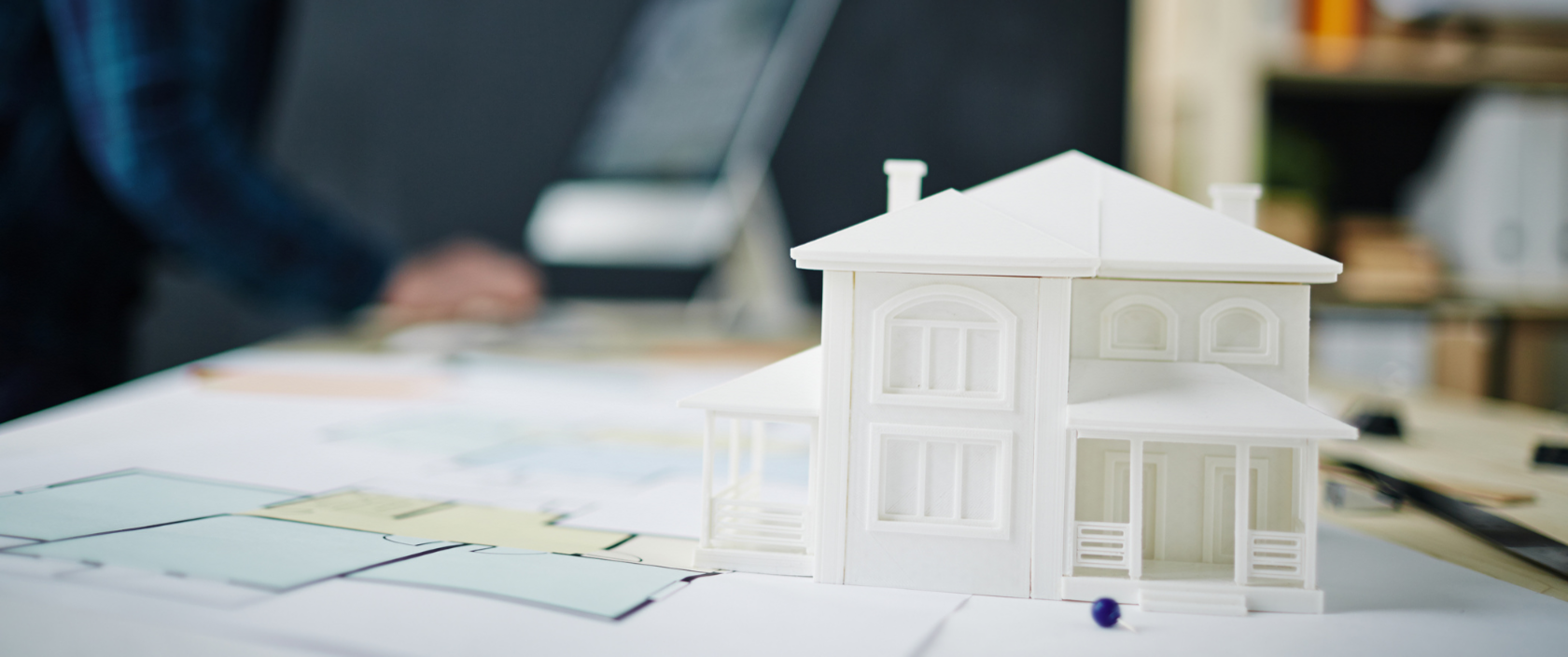 Miniaturhaus steht auf Bauplänen auf einem Schreibtisch, Mann arbeitet an Computer im Hintergrund