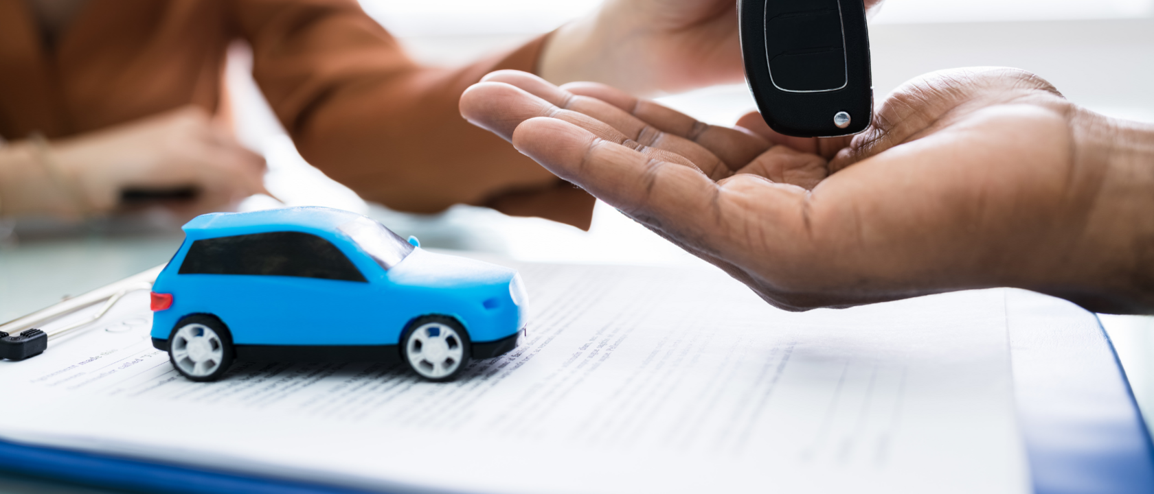 Miniaturauto steht auf Dokumenten, die auf einem Schreibtisch liegen, während ein Autoschlüssel übergeben wird.