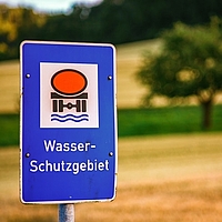 Schild Wasserschutzgebiet, Hintergrund unscharf mit Einzelbaum, Feld, Wiese und ganz hinten Wald