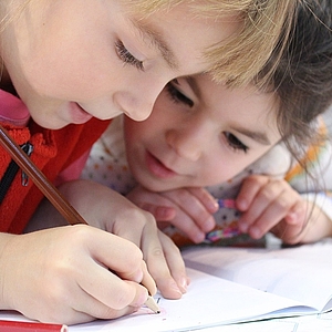 Zwei kleine Mädchen erledigen gemeinsam ihre Hausaufgabe. Die eine hält einen Stift und schreibt in ein Heft, die andere guckt interessiert zu