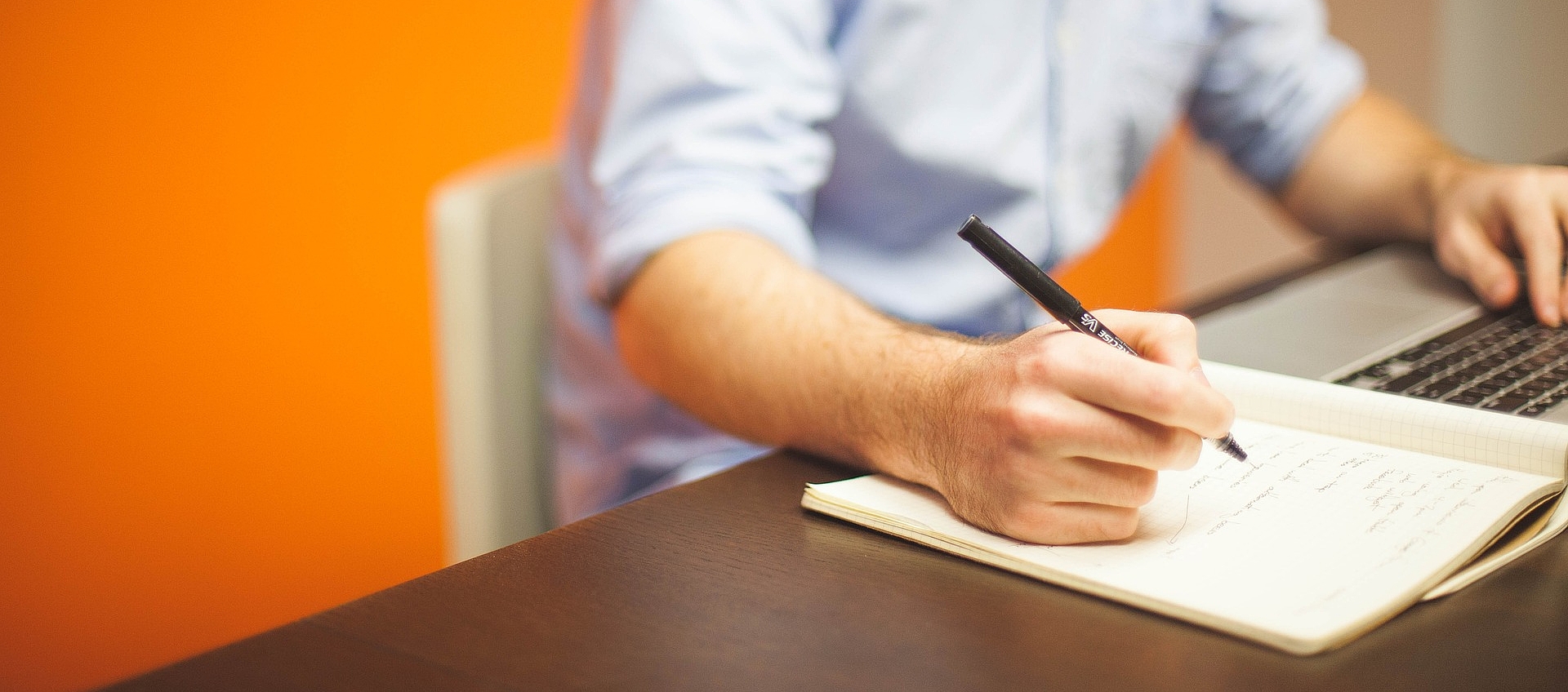Mann mit blauen Hemd sitzt an einem Schreibtisch vor einem Laptop, hält einen Stift in der Hand