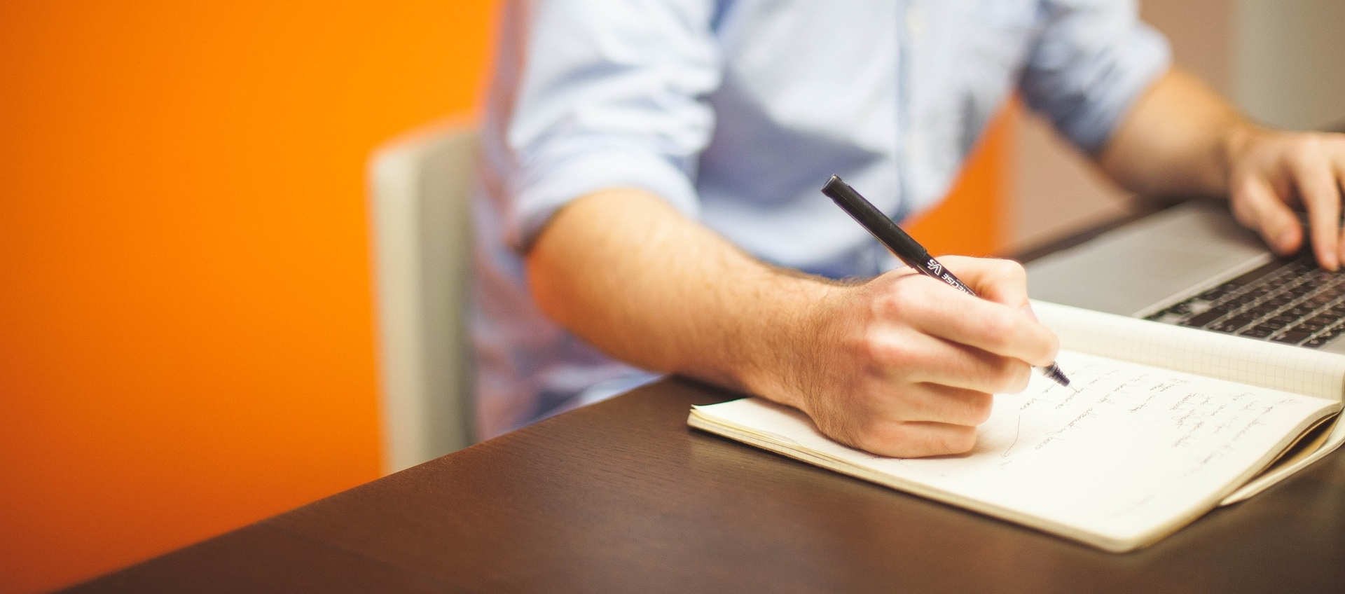 Mann mit blauen Hemd sitzt an einem Schreibtisch vor einem Laptop, hält einen Stift in der Hand