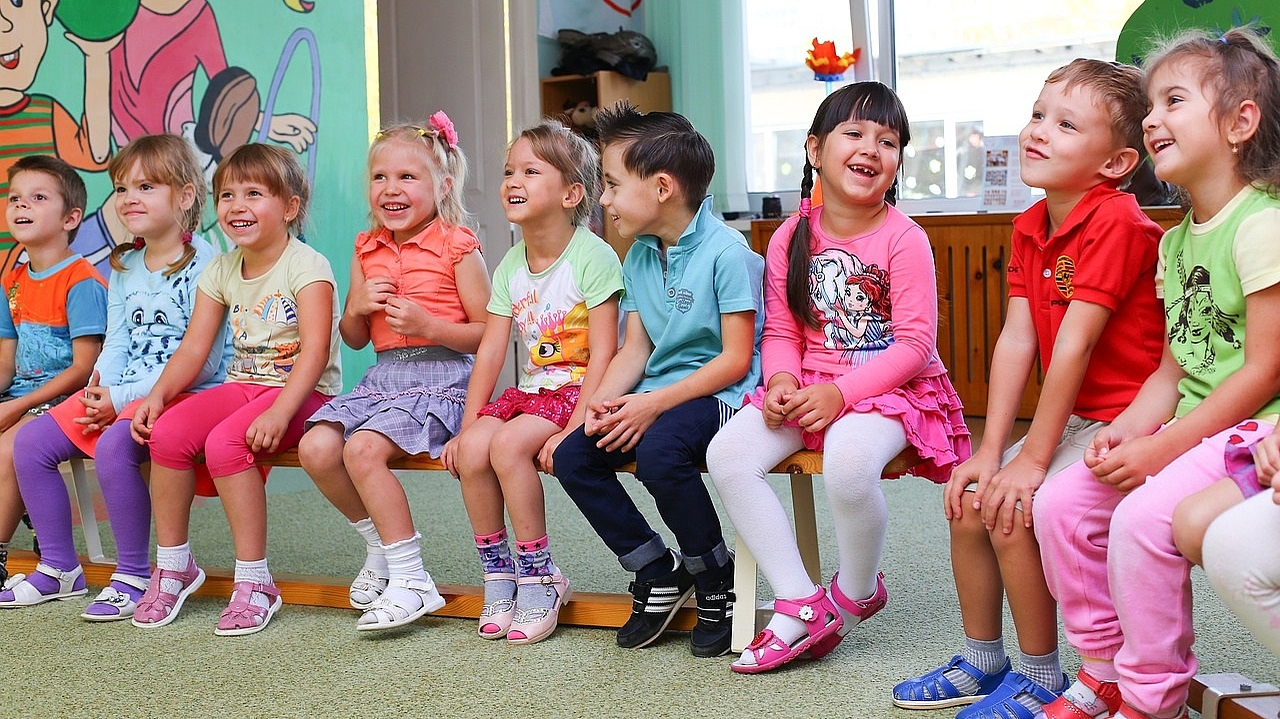 Eine Gruppe verschiedener Kinder, die im Kindergarten auf einer Bank sitzt, die Stimmung ist fröhlich, die Kinder lachen