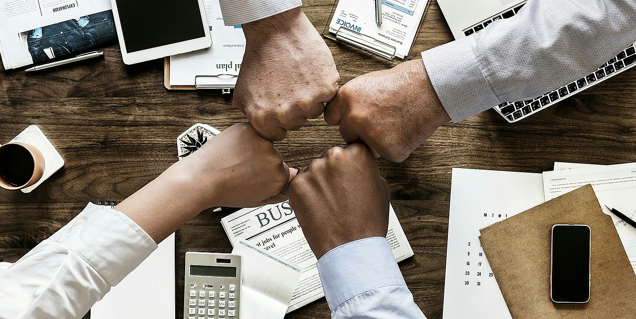 Vier Fäuste, die sich gemeinsam in der Mitte treffen. Über einem Tisch mit verschiedenen Arbeitsmaterialien.