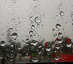 Senkrecht stehende Glasscheibe mit unscharfem grauen Hintergrund, rechts unten etwas rötlich mit Regentropfen, die teils als Linse für den Hintergrund wirken