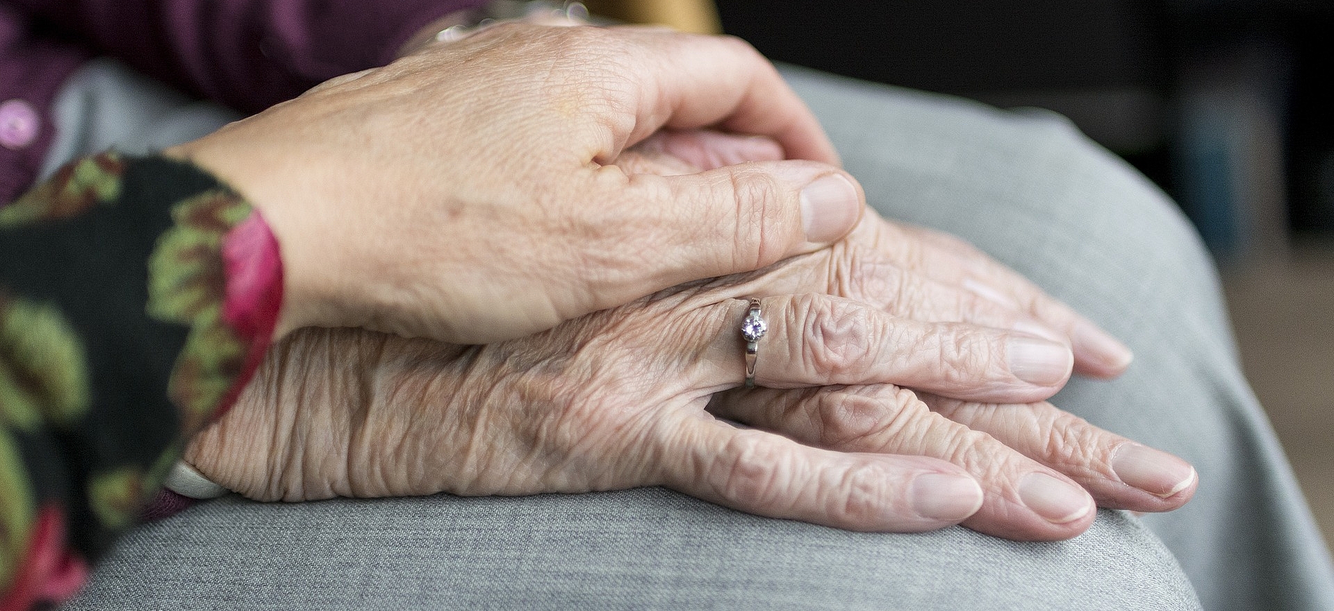 eine ältere Dame hat die Hände auf ihrem Schoß übereinander gefaltet liegen, die Hand einer weiteren Frau liegt beschützend über ihren Händen