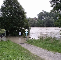 Kiesweg in einer Wiese am Inn, der in der Fortsetzung überschwemmt ist, zwei Schilder im Wasser, rechts der übervolle Inn, links, rechts und im Hintergrund Bäume