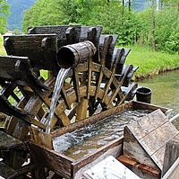 Holzmühlrad mit Wasserschöpfkrug Altes Holzmühlenrad mit Wasserschöpfer am Bach, im Hintergrund Wiese mit Baumgruppe rechts und Siedlung links