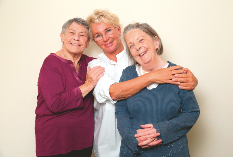 Drei ältere Frauen bzw. Seniorinnen umarmen sich und lächeln in die Kamera