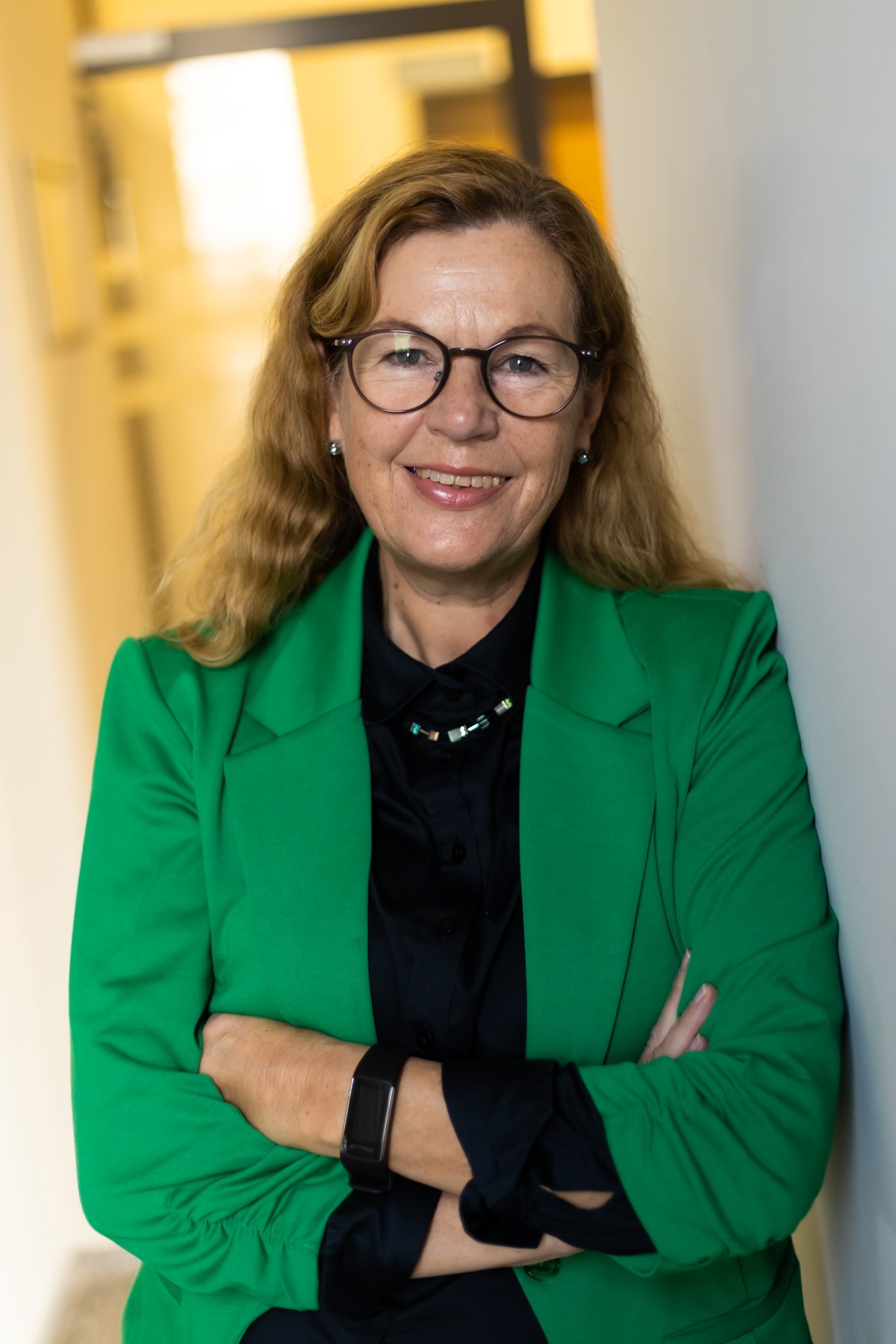 Frau mit grünem Jacket und Brille lehnt an einer Wand und lächelt