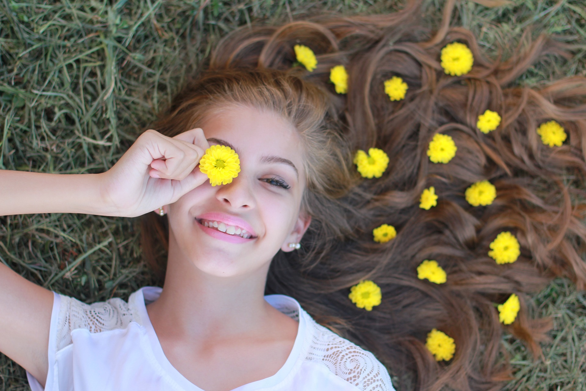 Ein Mädchen mit langen hellbraunen Haaren liegt auf einer grünen Wiese und lacht, vor ihr rechtes Auge hält sie eine gelbe Blüte, in ihren Haaren liegen weitere gelbe Blüten