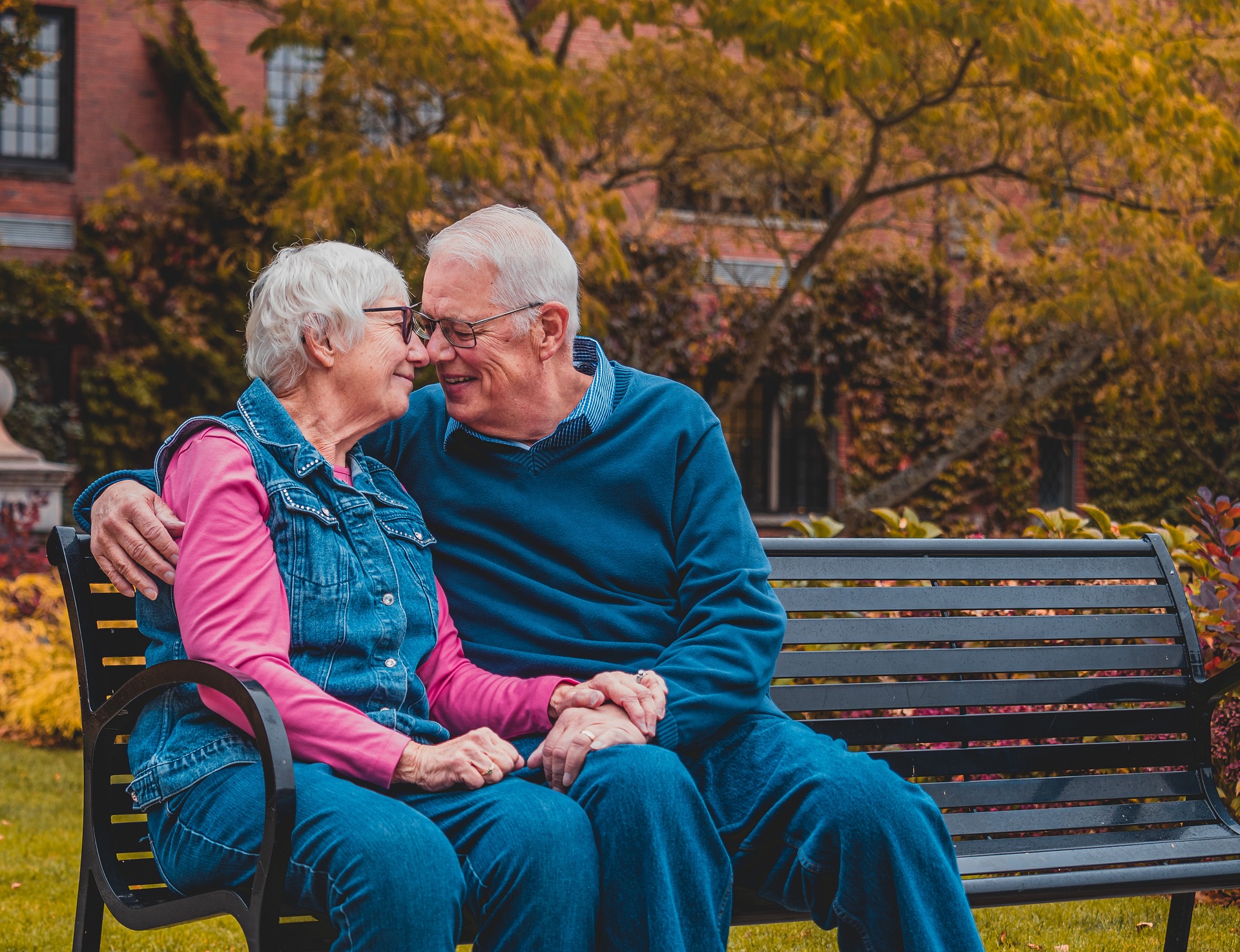 Älteres Ehepaar sitzt gemeinsam auf einer Parkbank, er hat den Arm um sie gelegt, sie halten Händchen und sehen sich verliebt an