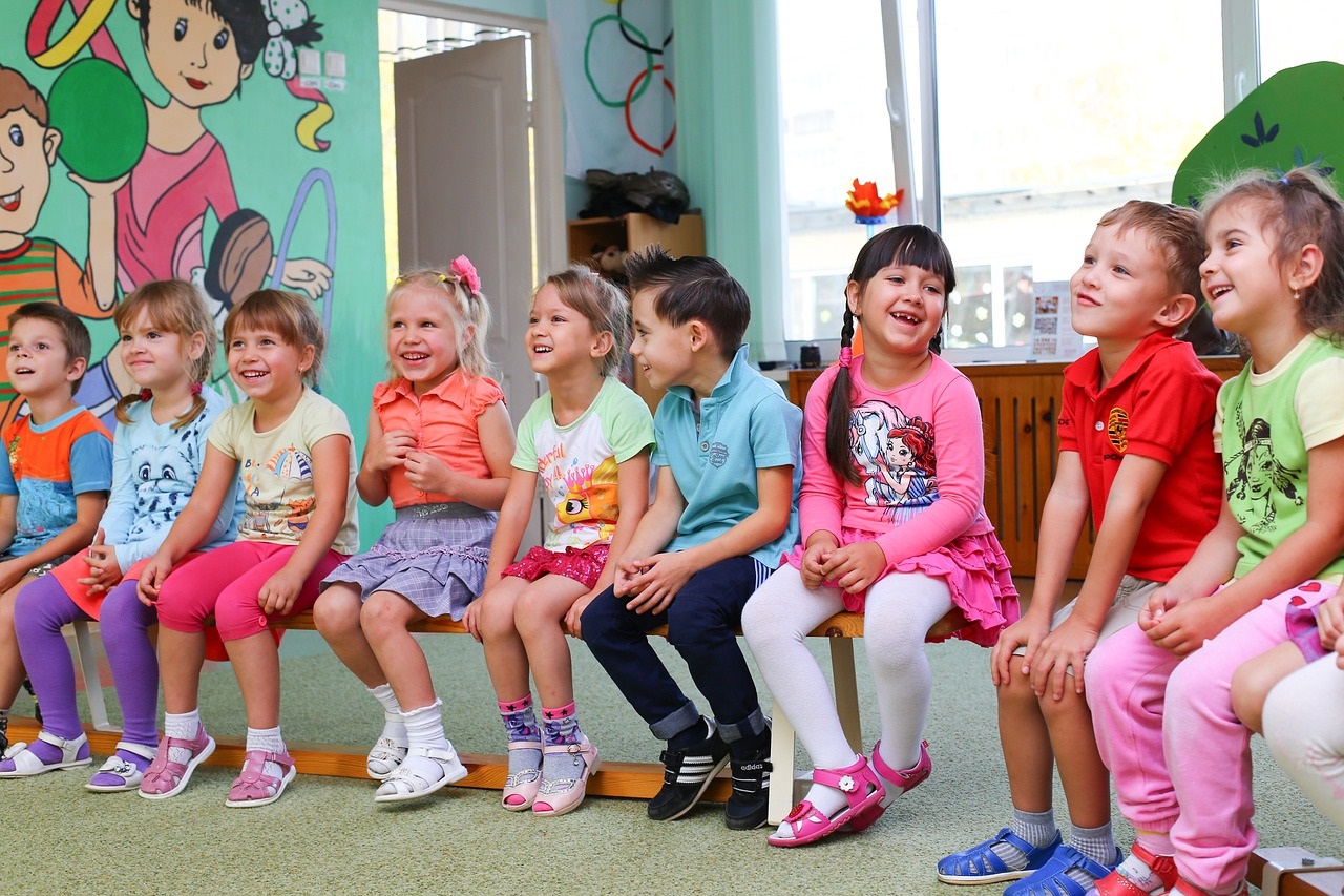 Eine Gruppe verschiedener Kinder, die im Kindergarten auf einer Bank sitzt, die Stimmung ist fröhlich, die Kinder lachen