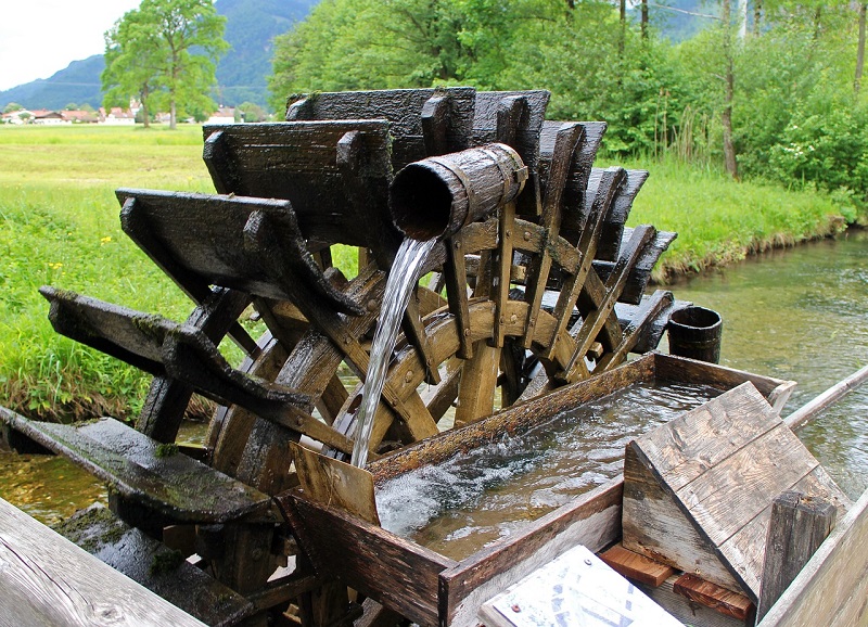 Altes Holzmühlenrad mit Wasserschöpfer am Bach, im Hintergrund Wiese mit Baumgruppe rechts und Siedlung links